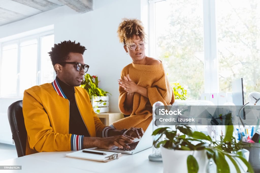 Colleagues working together on a new project Two business colleagues working together on a new project. African american businessman working on laptop with female coworker sitting on desk and helping him. 30-34 Years Stock Photo