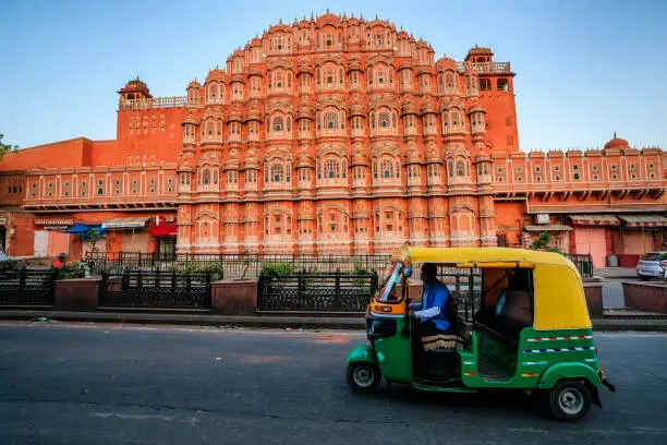 Indian man drives auto rickshaw (tuk-tuk) on streets of Rajasthan, India. Hawa Mahal (The Wind Palace) on the background.