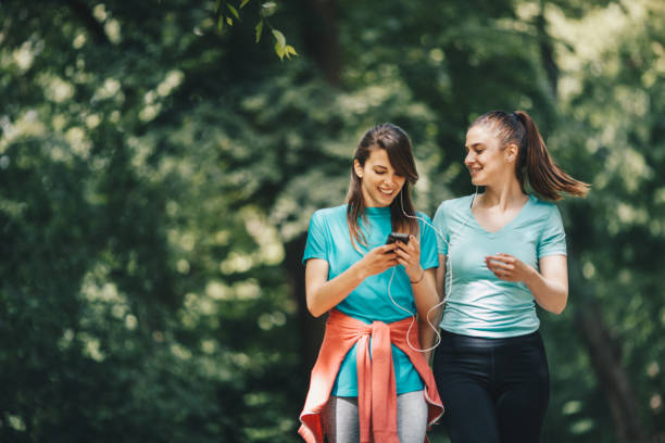 dwóch przyjaciół spacerujących po parku, słuchających muzyki ze smartfona i rozmawiających. - hiking young women outdoors t shirt zdjęcia i obrazy z banku zdjęć