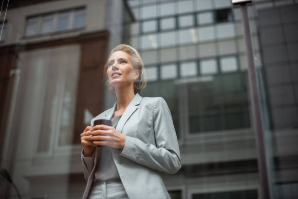 German businesswoman 40-year-old satisfied caucasian businesswoman is looking through the window from the bright office. She is holding a cup of coffee in hands. georgijevic frankfurt stock pictures, royalty-free photos & images