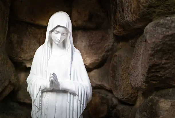On the corner of a small cathedral, there is a statue of the Virgin Mary praying in a stone-covered place.