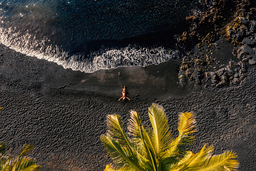 Drone shot aerial view of Young woman relaxing on tropical black sand beach in Hawaii . People travel luxury vacations destinations concept