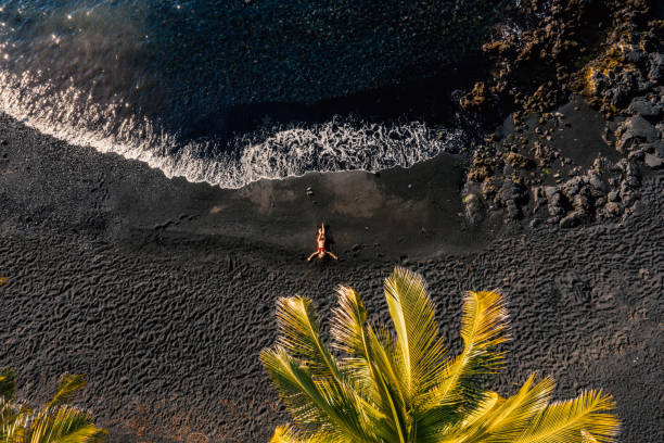 Drone view of Young woman lying on black sand beach Drone shot aerial view of Young woman relaxing on tropical black sand beach in Hawaii . People travel luxury vacations destinations concept black sand stock pictures, royalty-free photos & images