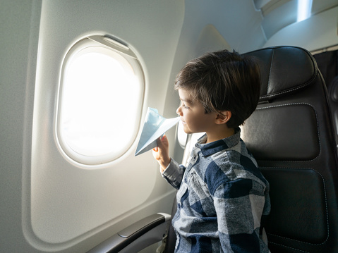 Cute little boy on an airplane looking at window view holding a paper airplane smiling