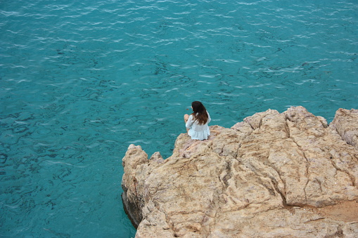 figure from the back, in the distance, of a girl unrecognizable who scrutinizes the sea, the infinite, sitting alone on a rocky cliff on the ocean water in a white dress in ibiza, spain