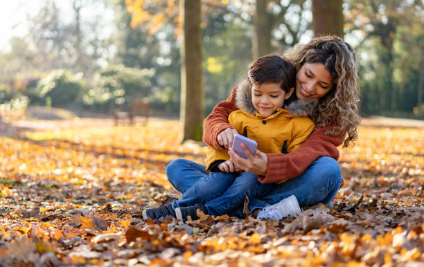 mutter und sohn genießen einen tag im park im herbst sitzen auf dem boden spielen mit dem smartphone - telephone nature mobile phone autumn stock-fotos und bilder