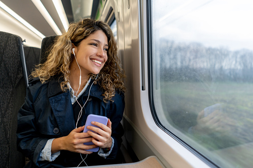 Happy woman traveling by train listening music with headphones while looking at the window view smiling