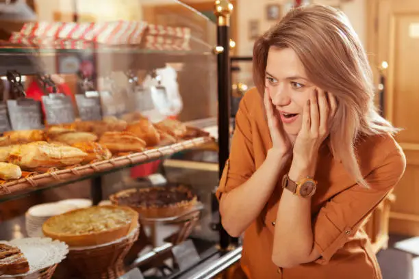 Excited mature woman shopping for pastry at local bakery, looking overwhelmed