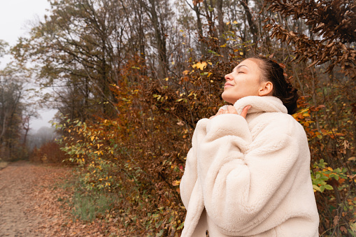 Attractive brunette young female wrapped in white coat stands among the colorful forest with closed eyes, autumn time