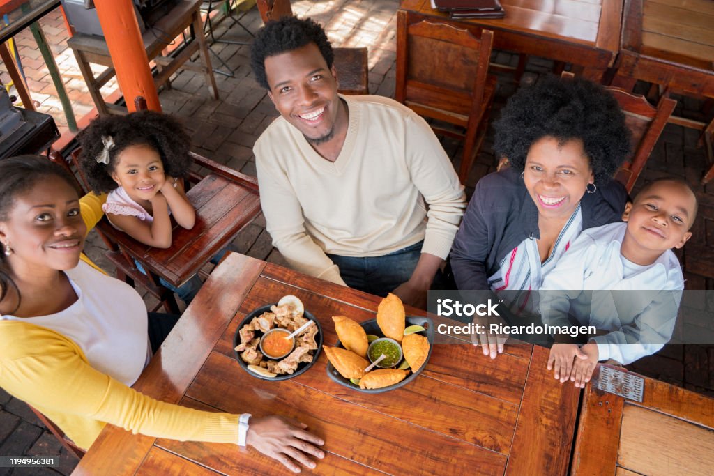 the afro family begins to eat the empanadas and the pork rinds they bring to the restaurant table The Afro family of mom, dad and daughter smile because the food arrived and they are delicious empanadas and chicharrones that they bring to the table of the restaurant of typical Colombian foodThe Afro family of mom, dad, grandmother and children smile because the food arrived and they are delicious empanadas and chicharrones that they bring to the restaurant table of typical Colombian food. Colombia Stock Photo