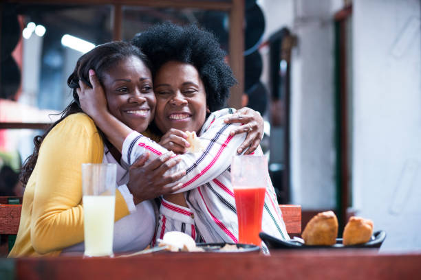 mom and daughter afro hair latin women sitting at typical restaurant table look each other in the eyes and smile at the table we see glasses of juices and empanadas - couple restaurant day south america imagens e fotografias de stock