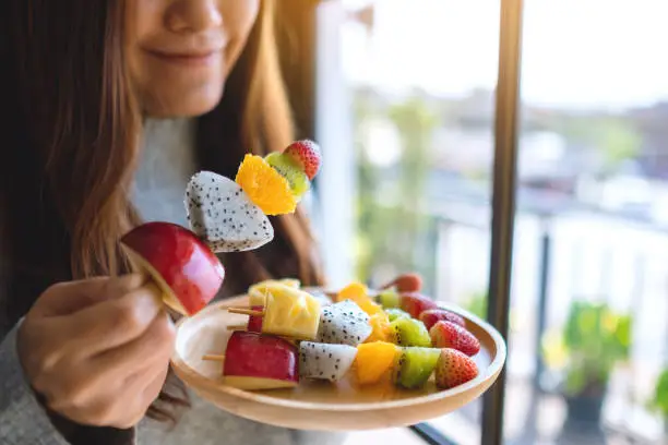 Photo of an asian woman holding and eating a fresh mixed fruits on skewers