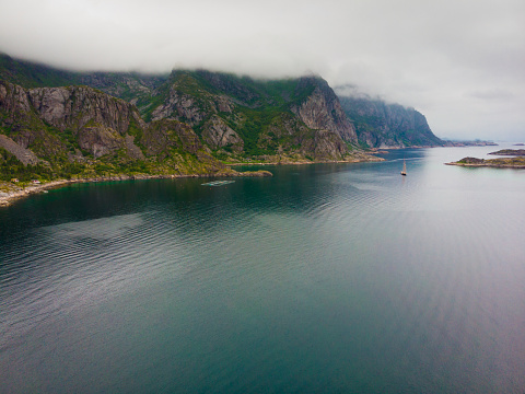 Sea landscape with yacht boat and stone islets among the waters of fjord Vjestfjord, Lofoten islands, Henningsvaer region, Norway. Hazy day, overcast weather.