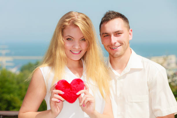 Love, romantic relationships concept. Man and blonde woman walking sightseeing on sunny day holding heart shape pillow enjoying romance, happy being together. City in background
