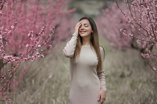 A woman in a long tight dress walks down a row between peach trees with a blossom tree.