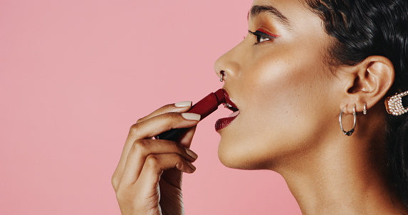 Studio shot of a beautiful young woman applying lipstick while standing against a pink background