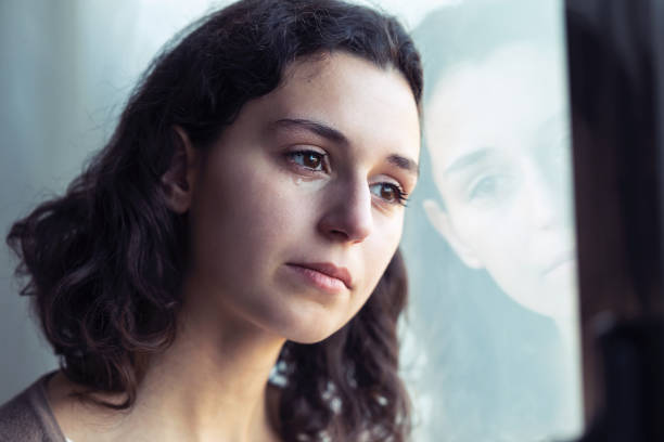 istock Sad young woman crying while looking through the window at home. 1195036200