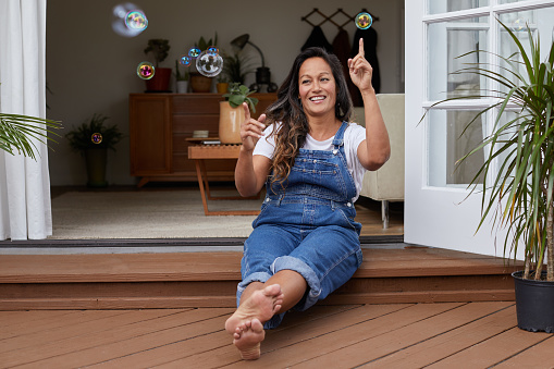 Mature woman sitting on her living room patio and laughing while popping soap bubbles