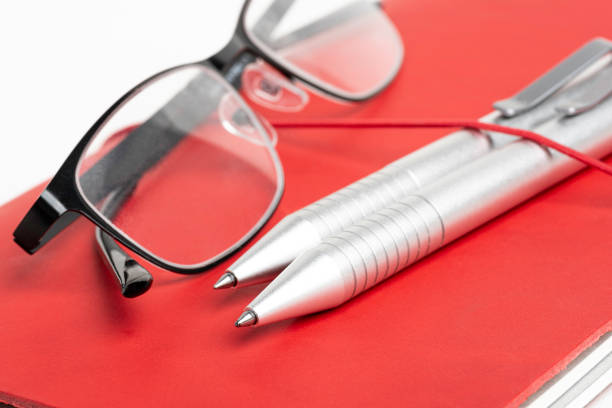A journal with red leather cover and elastic band securing two silver ballpoint pens and a pair of reading glasses.