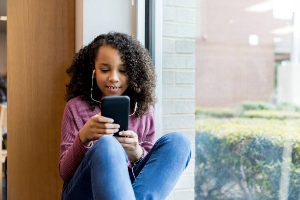 istock Girl sitting on windowsill in library texts with smart phone 1195063484