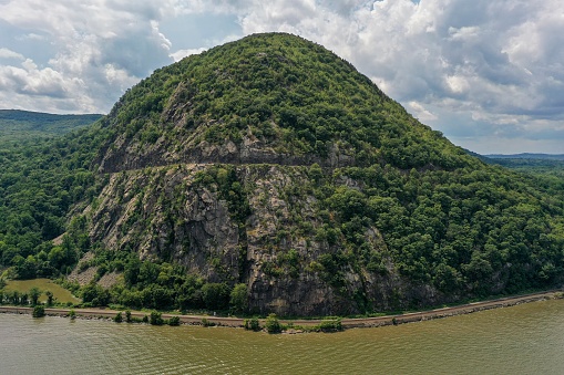 An daytime aerial view of a riverside mountain that bulges out of the green earth.