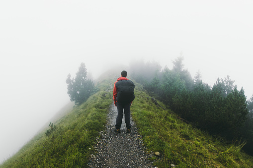 Young man tourist with backpack in red jacket enjoying his hiking trip through beautiful foggy footpath in the mountains of Appenzell region in Switzerland