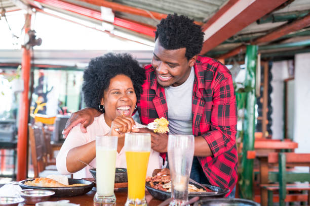 Latin woman with dark skin and afro hair of approximately 50 years dressed casually is sitting at a restaurant table where she has lunch with her family typical Colombian food while her son-in-law Latin man with brown skin and afro hair looks at her mujer latina con piel oscura y pelo afro de aproximadamente 50 años vestida casualmente está sentada en una mesa de restaurante donde almorza con su familia comida típica colombiana mientras su yerno hombre latino con piel morena y pelo afro la mira
