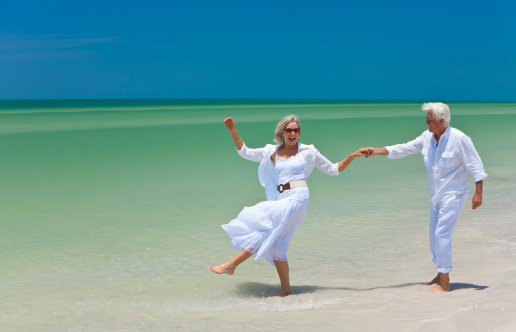 Happy senior man and woman couple dancing and holding hands on a deserted tropical beach with bright clear blue sky