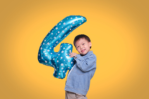 Adorable Asian four year old boy celebrating his birthday holding number 4 blue balloon on orange colored background with clipping path, Soft & Selective focus
