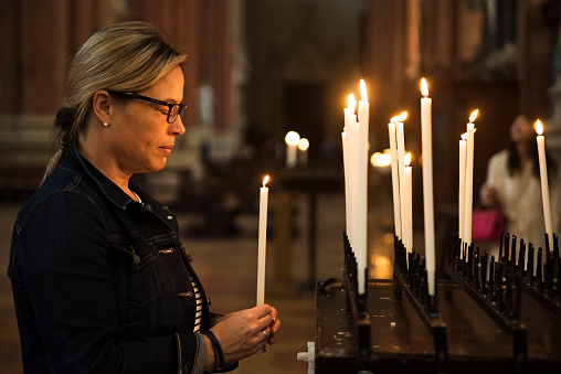 Everyday life of freshly expatriate mature woman discovering Italy. Here she is taking time to reflect on her journey and make a wish lighting some candles in a church. Horizontal waist up outdoors shot.