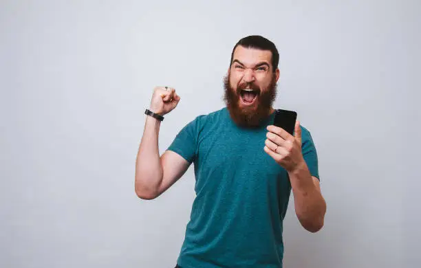 Photo of Bearded man, winner eith smartphone in hand screaming on white background.