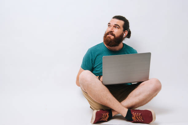 Happy bearded hipster man sitting on the floor with laptop computer and looking away