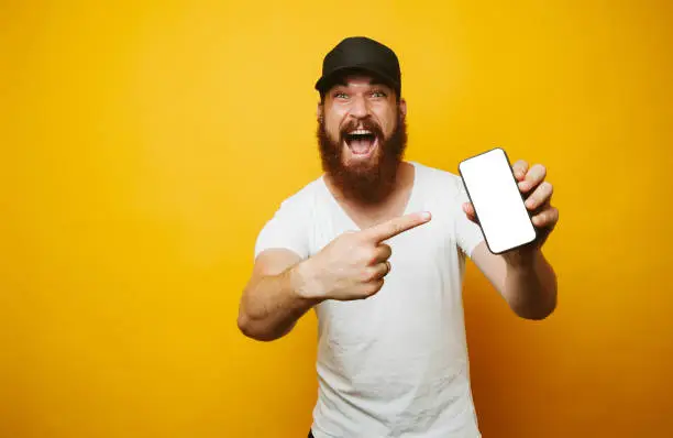 Photo of Amazed young bearded man in white t-shirt pointing at phone