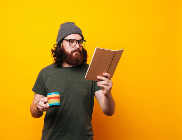 brodaty hipster w zielonej koszulce w okularach i kapeluszu, czytając książkę i trzymając filiżankę kawy - sitting young men book isolated on white zdjęcia i obrazy z banku zdjęć