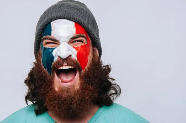 Photo of Face of young happy bearded man painted with flag of France. Football or soccer team fan, sport event, faceart and patriotism concept. Studio shot at gray background, copy space