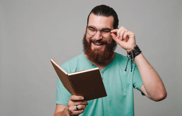 Happy young bearded man reading a book and smiling over grey background stock photo