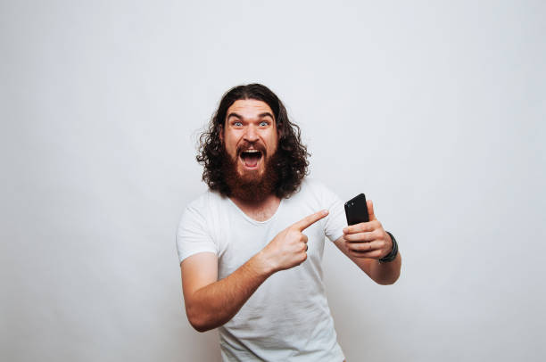 istock Surprised young bearded man pointing at phone over white background 1195123566