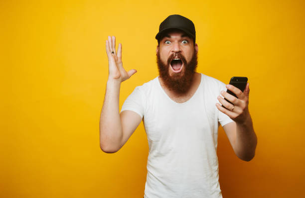 istock Shocked bearded man holding phone on yellow background. 1195123772