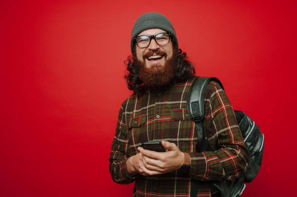 Portrait of happy man with beard wearing a backpack and holding phone and looking at the camera over red background stock photo