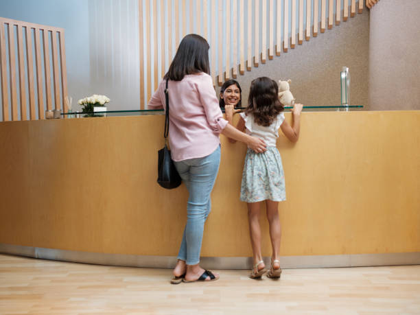 Latin mother and girl on tiptoes talking to receptionist A latin mother and a girl on tiptoes talking to the receptionist at a office's desk. medical office lobby stock pictures, royalty-free photos & images