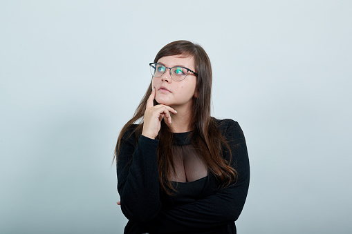 Boring young caucasian woman posing isolated on gray background in studio wearing casual dress, keeping hand on chin, thinkink about issue