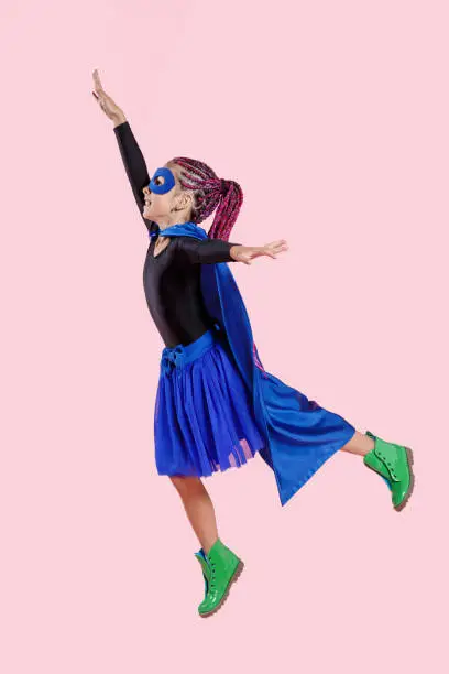 Photo of Vertical view. Little girl plays superhero. Kid on the background of bright pink wall, wear in colorful clothes.