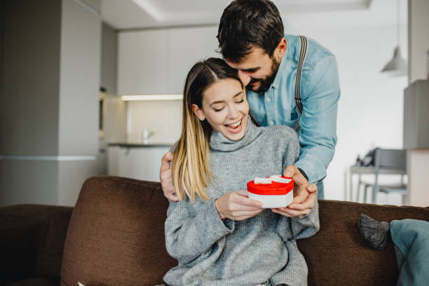 Young romantic couple holding present Young couple is at home. They are sitting on a couch, head to head and holding a present valentines present stock pictures, royalty-free photos & images