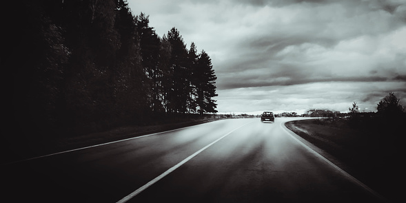 Moody black and white landscape with dark storm clouds, forest, car and road out of horizon. Beautiful nature background and wallpaper