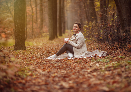 Art portrait. Young attractive woman sits on a plaid and drinks tea in the autumn forest