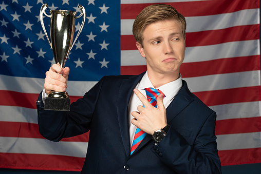 man imitating politician pointing with finger at golden goblet on american flag background