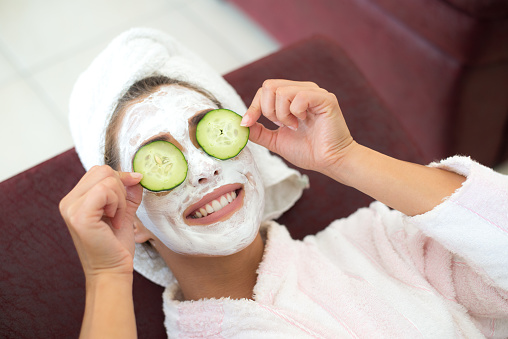 Happy woman putting cucumber facial mask on her face while lying on the sofa.
