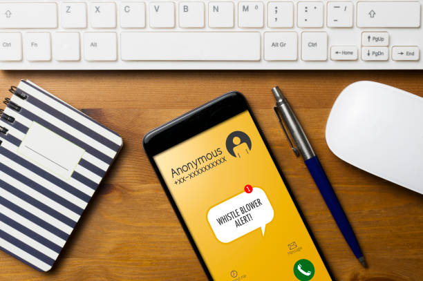 Whistleblowing concept: smartphone on wooden table with a computer keyboard, an agenda a pen and a mouse, show a whistle blower alert on an anonymous call