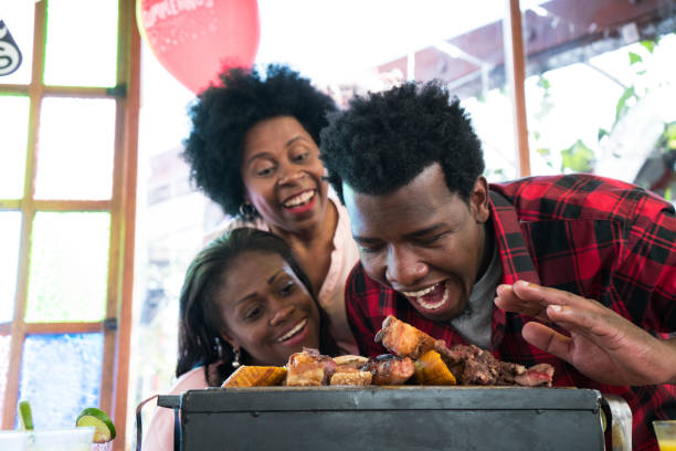 lateinischer mann mit afro-haaren und brauner haut, ca. 29 jahre alt, hat einen grill mit fleisch von chorizo-wurst vor sich, schaut in die kamera und öffnet seinen großen mund, während wir auf seine frau und schwiegermutter zurückblicken, die ihn beob - onionskin stock-fotos und bilder