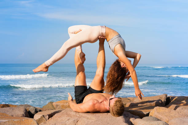 Fit young couple doing acro yoga exercise at sea beach Fit young couple doing acro yoga at spa retreat on sea beach. Active woman balancing on partner feet, stretching at acroyoga pose. Healthy lifestyle. People outdoor sport activity on family vacation. acroyoga stock pictures, royalty-free photos & images